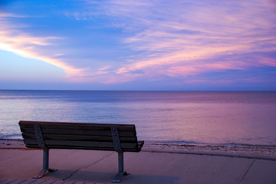 A park bench overlooks the calm waters of Long Island Sound as shades of pink, purple, and blue illuminate the evening sky following a passing storm on Long Island's North Fork. The peaceful waterfront scene captures the quiet beauty that makes the island's shoreline one of the best places in New York to experience a spectacular sunset. File photo: Diane Woodcheke, licensed.