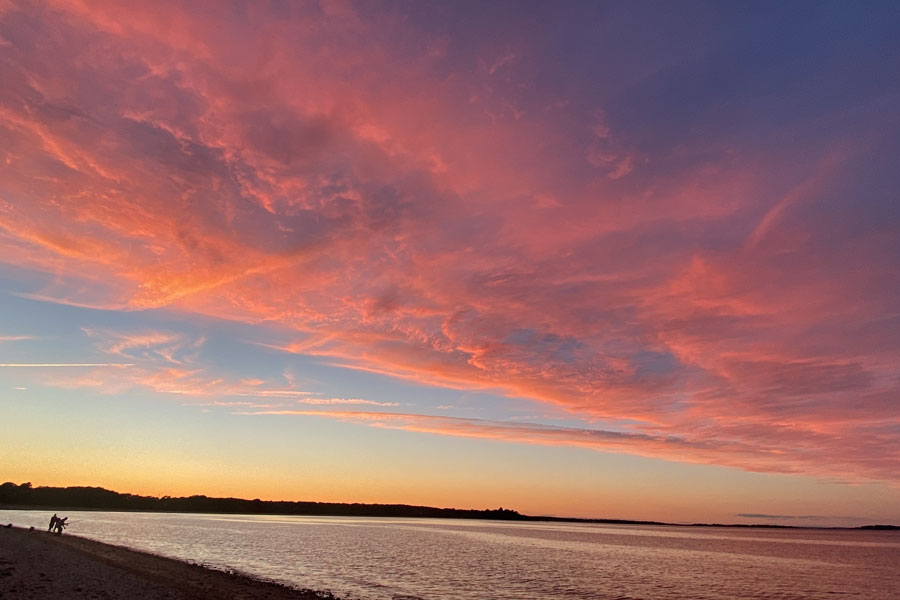 A vibrant sunset paints the sky above Shelter Island with dramatic shades of pink, orange, and purple as calm bay waters reflect the fading light. Two fishermen stand silhouetted along the shoreline, adding a peaceful sense of scale to the expansive waterfront scene. The colorful cloud formations and tranquil evening atmosphere capture the natural beauty that makes Shelter Island one of Long Island's most scenic places to end the day.