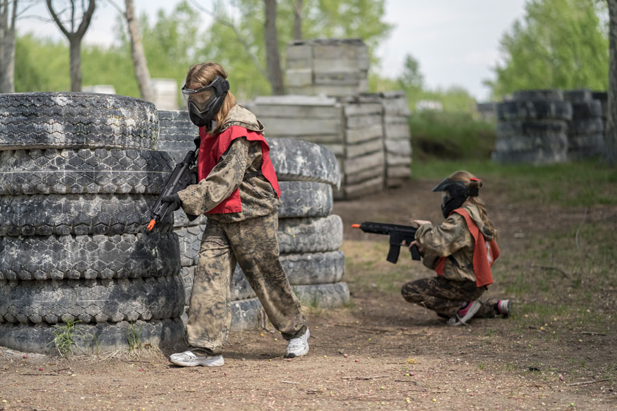 Two airsoft players move between tire and wooden barricades during an outdoor match, wearing protective masks and team vests while navigating the field. Airsoft facilities on Long Island offer tactical, scenario-based play for individuals, groups, parties, and experienced players.