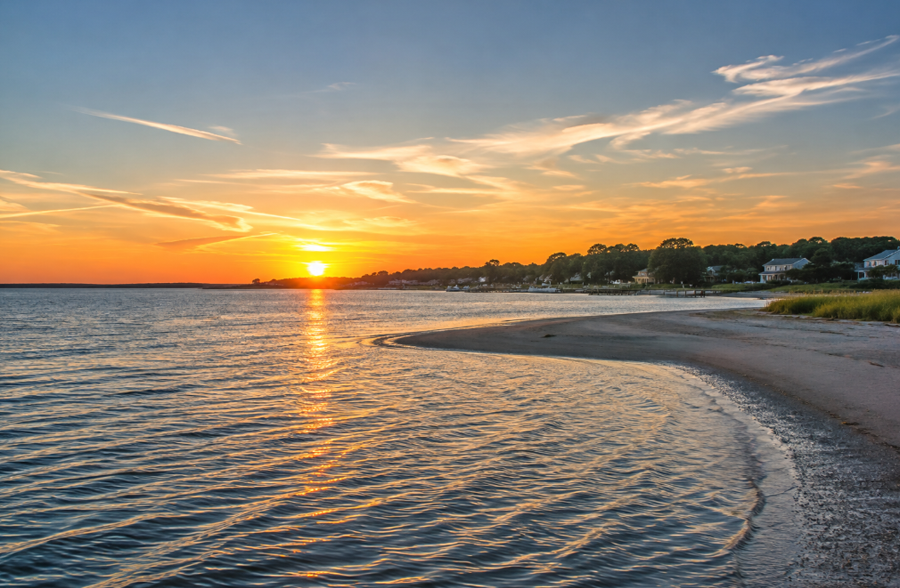 Sunset over Bellport Bay casts warm light across the calm shoreline and gently rippling water along Long Island’s South Shore. The scene reflects the bay’s quiet coastal character, where protected waters and sandy inlets meet the communities bordering eastern Great South Bay.