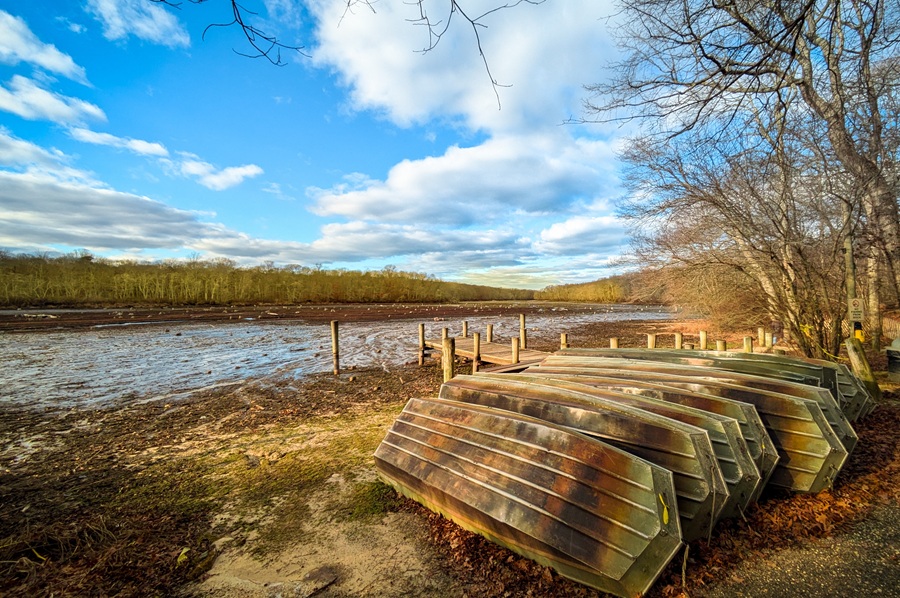 An autumn view of the drained Stump Pond at Blydenburgh County Park in Smithtown after damage to the New Mill Pond Dam caused the lake to empty. Stacked rowboats remain beside the exposed shoreline, while the former lakebed stretches beneath a bright blue sky and late-season trees.