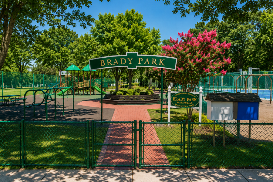 A sunny view of Brady Park in Massapequa Park, showing the park’s distinctive green entrance signage, fenced playground, colorful play equipment, sports courts, landscaped grounds and mature shade trees.