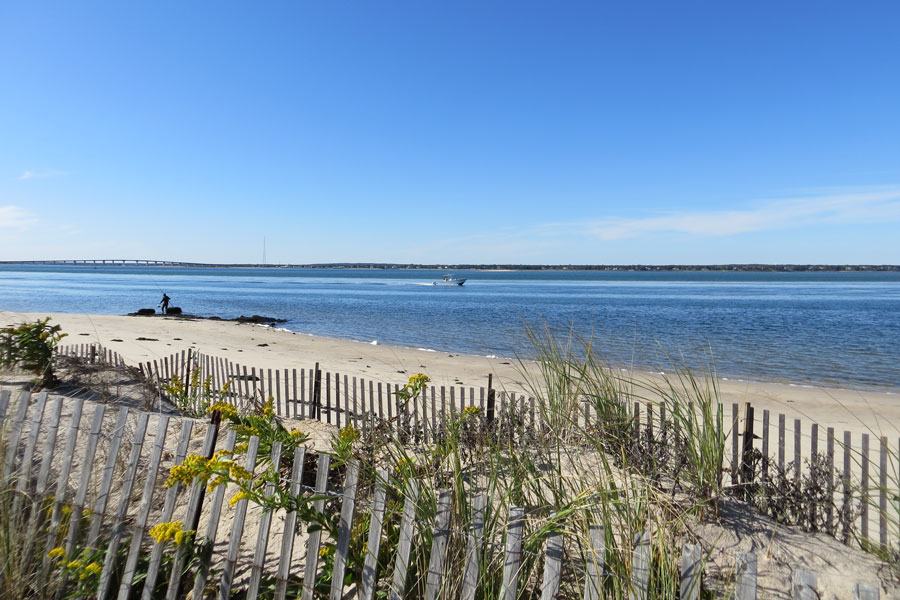 Clear blue skies stretch over Shinnecock Bay at Shinnecock East County Park in Southampton, Long Island. Low dunes and a quiet sandy shoreline frame the calm bay waters on the barrier beach east of Shinnecock Inlet. The park’s open setting provides a more rugged, undeveloped coastal landscape than a typical South Fork beach.
