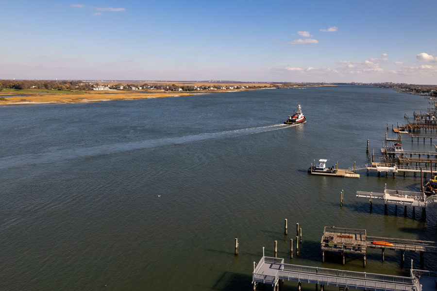 Aerial view of East Rockaway Inlet, with a tugboat moving through the broad channel between marsh-lined shoreline and waterfront docks. The scene highlights the inlet’s role as a navigable connection between the Atlantic Ocean, Hempstead Bay and the protected waterways of southwestern Nassau County.
