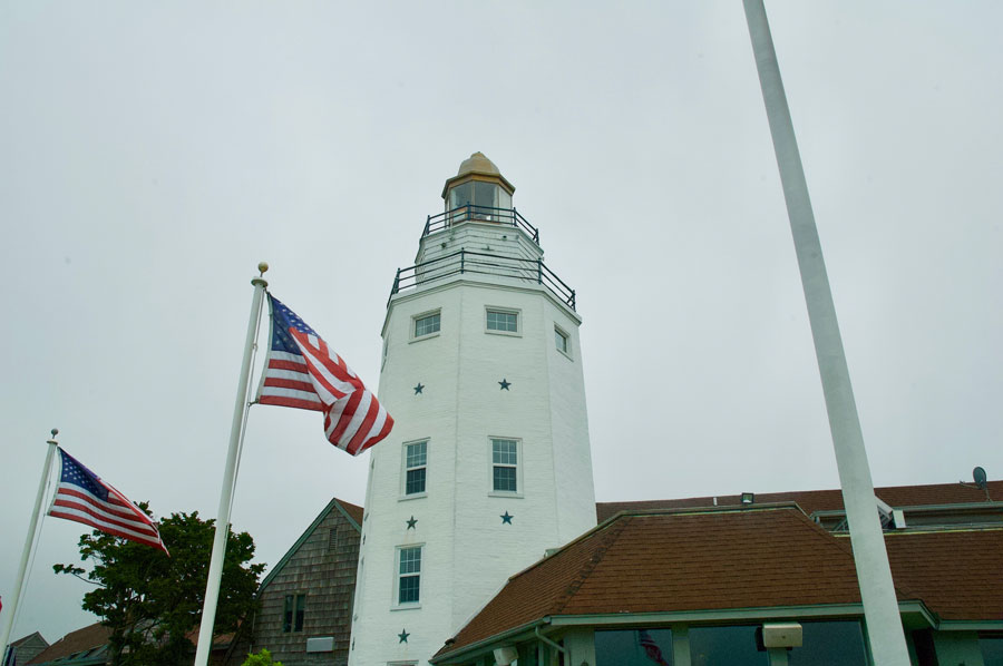 The lighthouse at the Montauk Yacht Club rises above the resort’s waterfront buildings, with American flags waving in the foreground. The distinctive private landmark resembles Montauk Point Lighthouse and overlooks Star Island in Lake Montauk.
