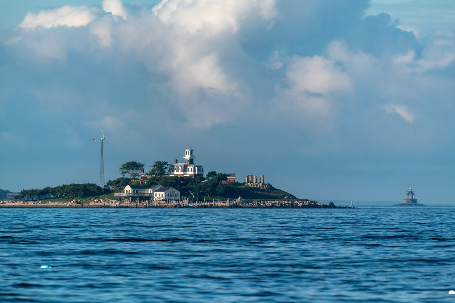 North Dumpling Island rises from Fishers Island Sound, with its historic lighthouse and private buildings visible along the shoreline. Race Rock Lighthouse appears in the distance to the right, marking the challenging waters of The Race.