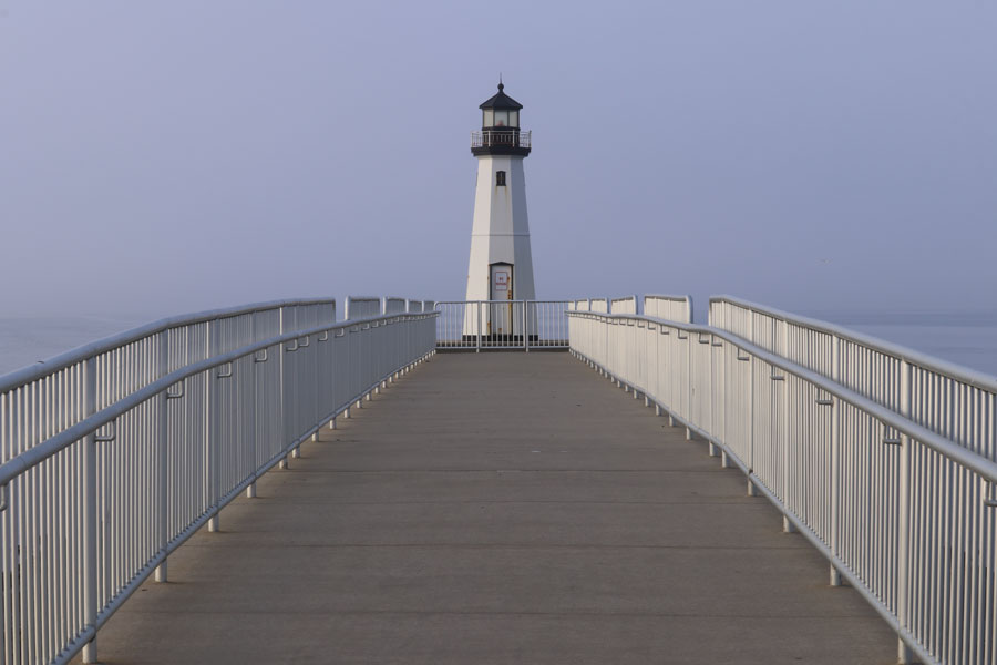 The Patchogue River Lighthouse stands at the end of a broad pedestrian jetty, surrounded by calm waters and morning haze at the entrance to the Patchogue River. The modern navigational light is part of the Sandspit Marina waterfront in Patchogue, Long Island.