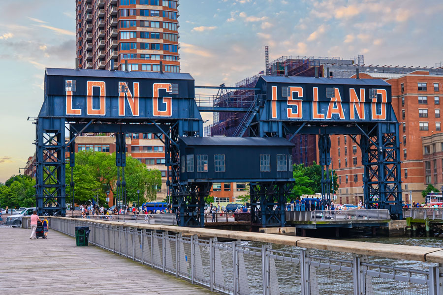 The iconic Long Island sign at Gantry Plaza State Park in Long Island City stands along the East River waterfront, with the neighborhood skyline rising behind it. The waterfront park is one of many places to explore across Long Island, from urban attractions and scenic parks to beaches, museums, farms, entertainment and outdoor recreation.