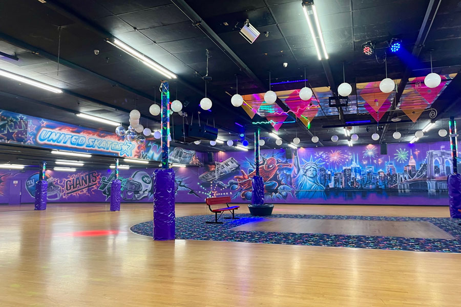 Inside United Skates of America in Seaford, Long Island, a colorful roller-skating rink features polished wood floors, neon lighting, suspended decorations, and a New York-themed mural overlooking the skating area. The family entertainment center hosts public skating sessions, parties, special events, and other roller-skating activities.