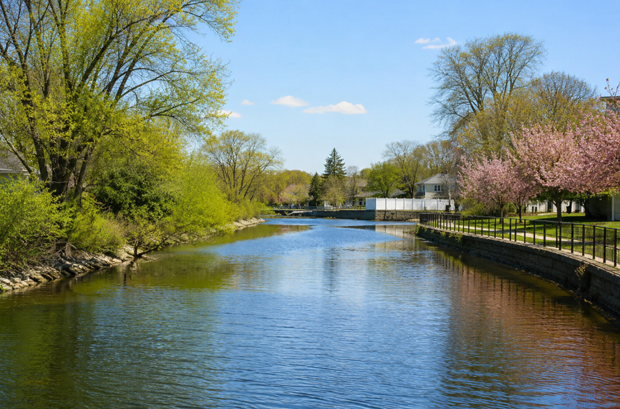 A peaceful view of Mill River shows calm blue water flowing past a landscaped shoreline lined with trees, fencing and nearby homes on a bright, clear day. The scene captures the river’s suburban waterfront character in southwestern Nassau County, where residential neighborhoods meet one of the South Shore’s connected tidal waterways.