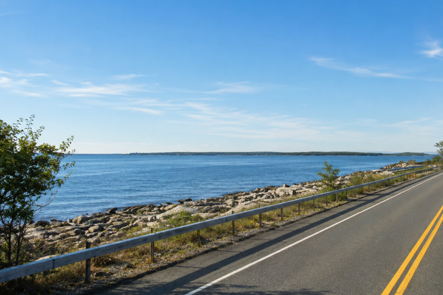 Scenic view of Orient Harbor from New York 25 in East Marion, where the shoreline road follows the calm blue waters of the eastern North Fork. The image captures the quiet coastal character of this historic harbor, framed by rocky shoreline, open water and wide sky.