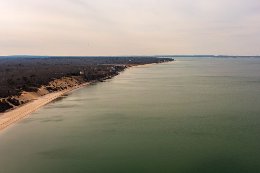 A high-angle aerial view of Meschutt Beach along the Peconic Bay shoreline in Hampton Bays, Long Island. The undeveloped beach, wooded shoreline and calm estuarine waters illustrate the coastal landscape near the Peconic River system.