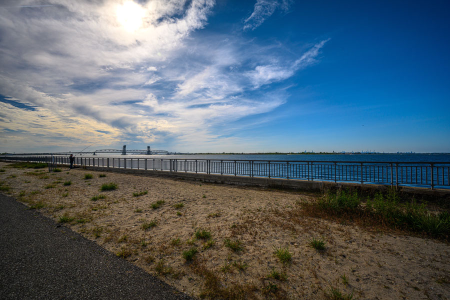 View across Rockaway Inlet from the shoreline, with a waterfront railing and fishing area overlooking the broad channel beneath a dramatic coastal sky. The Marine Parkway-Gil Hodges Memorial Bridge is visible in the distance, marking the entrance between the Rockaway Peninsula and Brooklyn as the inlet opens into Jamaica Bay.