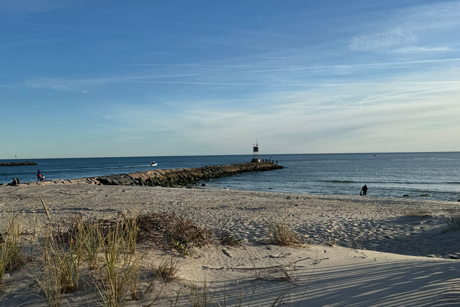 View across Shinnecock Inlet from the sandy shoreline, with the rock jetty extending into the Atlantic Ocean beneath a clear coastal sky. The inlet provides a vital passage between Shinnecock Bay and the open ocean and remains an important location for fishing, boating and maritime navigation.
