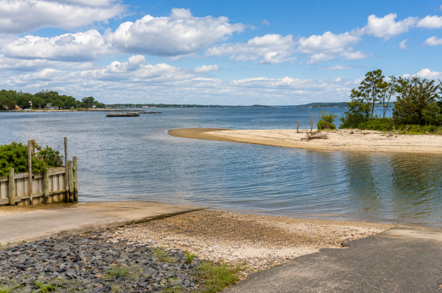 A scenic view across Southold Bay from the end of Pine Neck Road in Southold, with calm blue water opening toward the wider bay beneath a bright summer sky. A narrow sandy point, shoreline vegetation, small docks and distant waterfront homes highlight the quiet, sheltered character of this North Fork coastal setting.