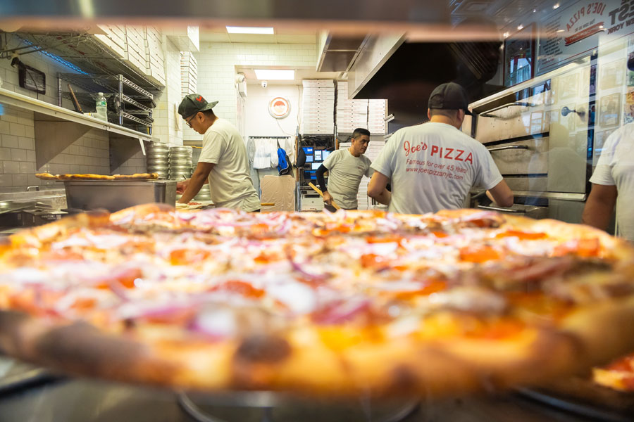 A freshly baked pizza sits on the counter as kitchen staff prepare additional pies inside a busy pizzeria. The scene captures the fast-paced atmosphere of a traditional New York-style pizza shop.