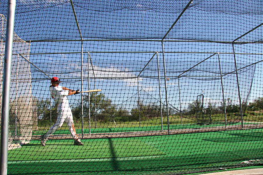 A batter takes a swing inside an outdoor batting cage, surrounded by protective netting and artificial turf under a clear blue sky. Batting ranges like this give baseball and softball players a convenient place to practice their timing, mechanics, and hitting throughout Long Island.