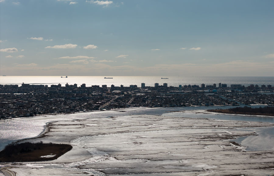Aerial view across the frozen marshes of Hempstead Bay toward Oceanside and the Atlantic Ocean, with cargo ships visible along the distant horizon. The winter scene highlights the broad tidal wetlands and protected waterways that define this part of Nassau County’s South Shore.