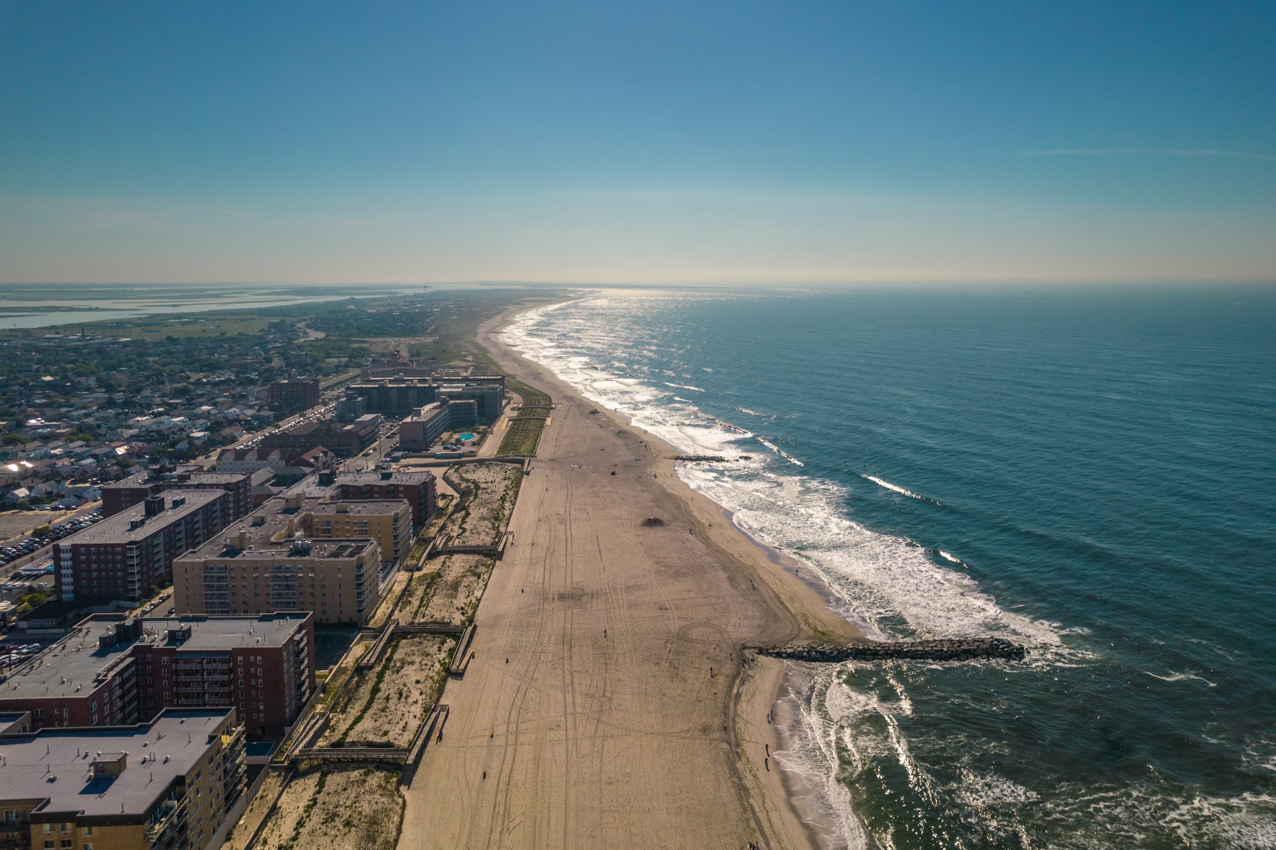 An aerial view looks west along the oceanfront in Long Beach, New York, with the Atlantic Ocean stretching beside the city’s wide sandy shoreline. Residential buildings, beachfront developments, jetties, and the barrier island landscape are visible beneath a clear blue sky.