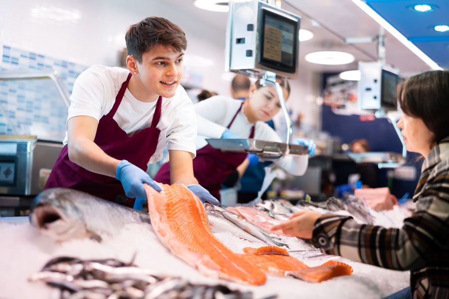 A seafood market employee displays a fresh salmon fillet to a customer at the counter. Fish and shellfish are arranged on ice as workers prepare orders behind the display.
