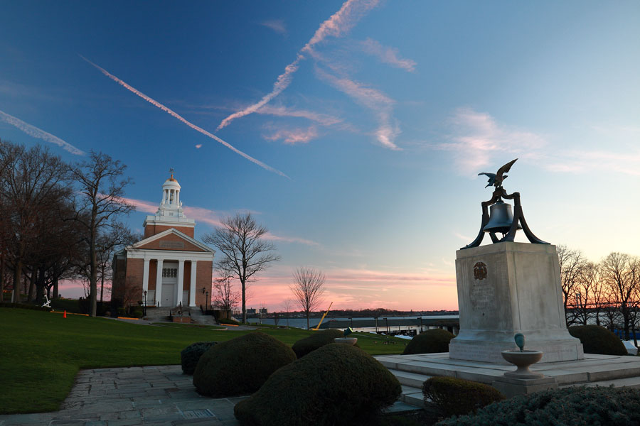 The United States Merchant Marine Academy campus in Kings Point at sunset, with the Mariners’ Memorial Chapel overlooking the waterfront and the Memorial Bell in the foreground. The federal service academy has trained merchant marine officers on Long Island’s Great Neck Peninsula since World War II. 