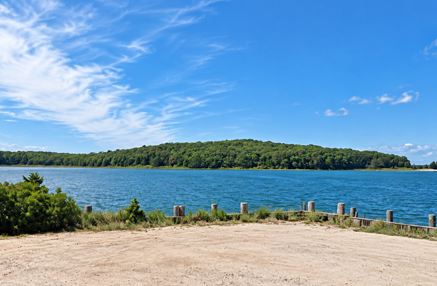 Give me a caption for this photo. A view across the waters of Northwest Harbor toward the densely wooded landscape of Barcelona Neck in East Hampton. Seen from Northwest Landing Road, the protected peninsula forms part of the Linda A. Gronlund Memorial State Forest and preserves more than 500 acres of coastal forest, wetlands, shoreline, and trails on Long Island’s East End.