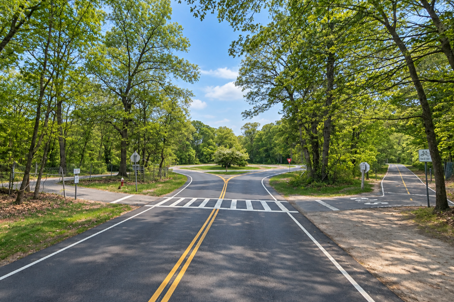 The Bethpage Bikeway crosses a park roadway within Bethpage State Park near Farmingdale, with paved sections of the multi-use trail continuing on both sides of the road. Surrounded by mature woodland, this portion of the route illustrates how the bikeway connects recreational areas throughout central Nassau County while periodically crossing local park roads.