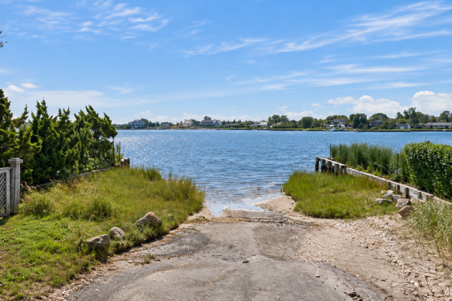 A peaceful view across Quantuck Bay from the end of Delafield Street in Westhampton Beach, where a narrow shoreline opening leads into the bay’s calm, protected waters. Marsh grasses and coastal vegetation frame the waterfront, with homes and docks visible along the opposite shore.