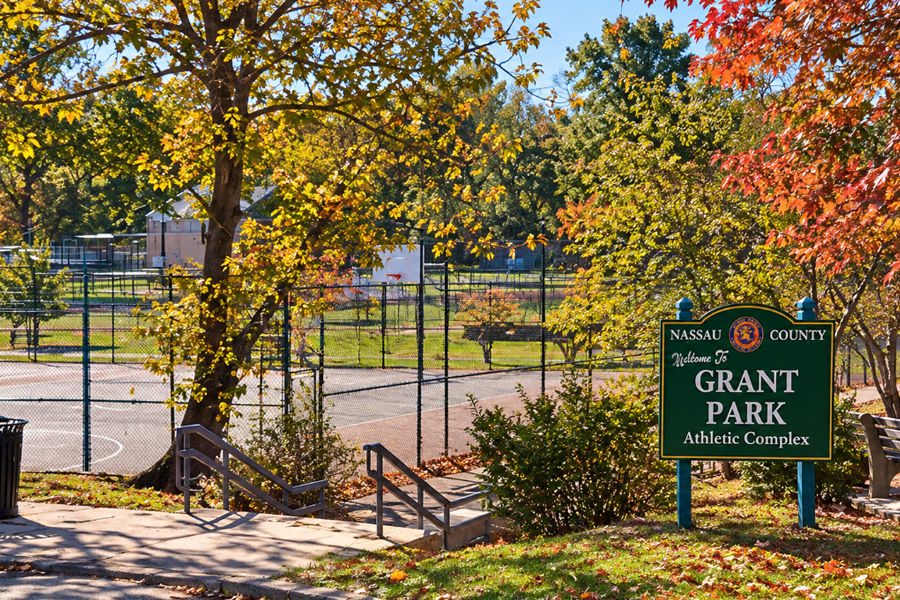 A recreated view of Grant Park in Hewlett, Nassau County, New York, featuring the park's athletic complex, basketball courts, walking paths, and distinctive entrance sign surrounded by colorful autumn foliage. The 35-acre Nassau County park offers sports facilities, playgrounds, a fishing lake, and year-round recreational opportunities.