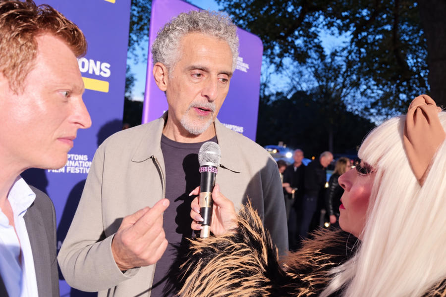 John Turturro speaks with Cognac Wellerlane as director Noah Segan looks on during red carpet coverage at the 2026 Hamptons International Film Festival. Turturro stars in Segan’s The Only Living Pickpocket in New York as an old-school hustler navigating a changing city. The interview was part of Wellerlane’s final day of festival coverage. AI-assisted recreation based on original video footage. Image credit: Cognac’s Corner Magazine and LongIslandGuide.com.