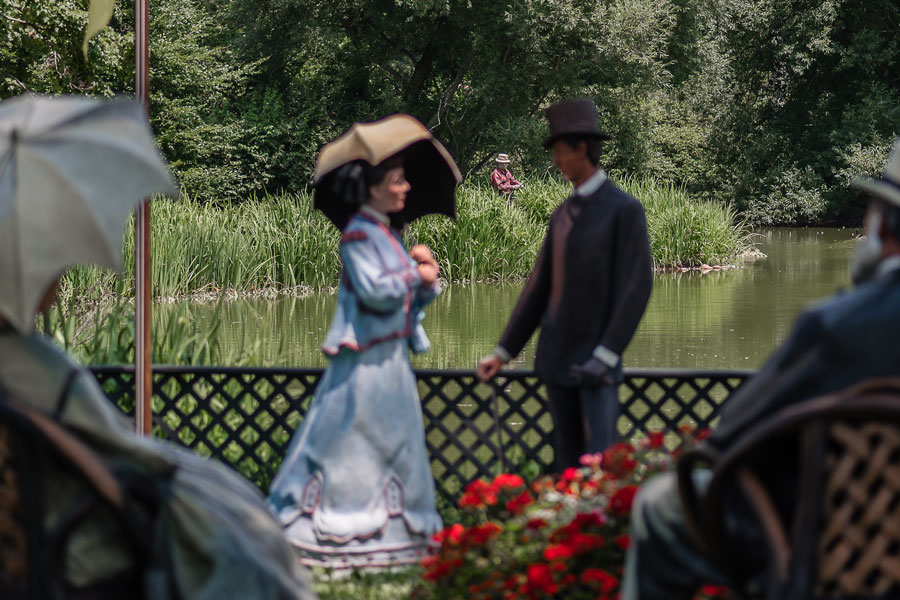 Life-size sculptures of elegantly dressed figures, including a woman with a parasol and a man in a top hat, create a garden-party scene beside a pond at Old Westbury Gardens in Old Westbury, New York. A fishing figure appears in the background of the Johnson sculpture exhibition, photographed on July 22, 2017, evoking the leisurely estate life associated with Long Island’s Gold Coast era. File photo: Jay Gao, licensed..