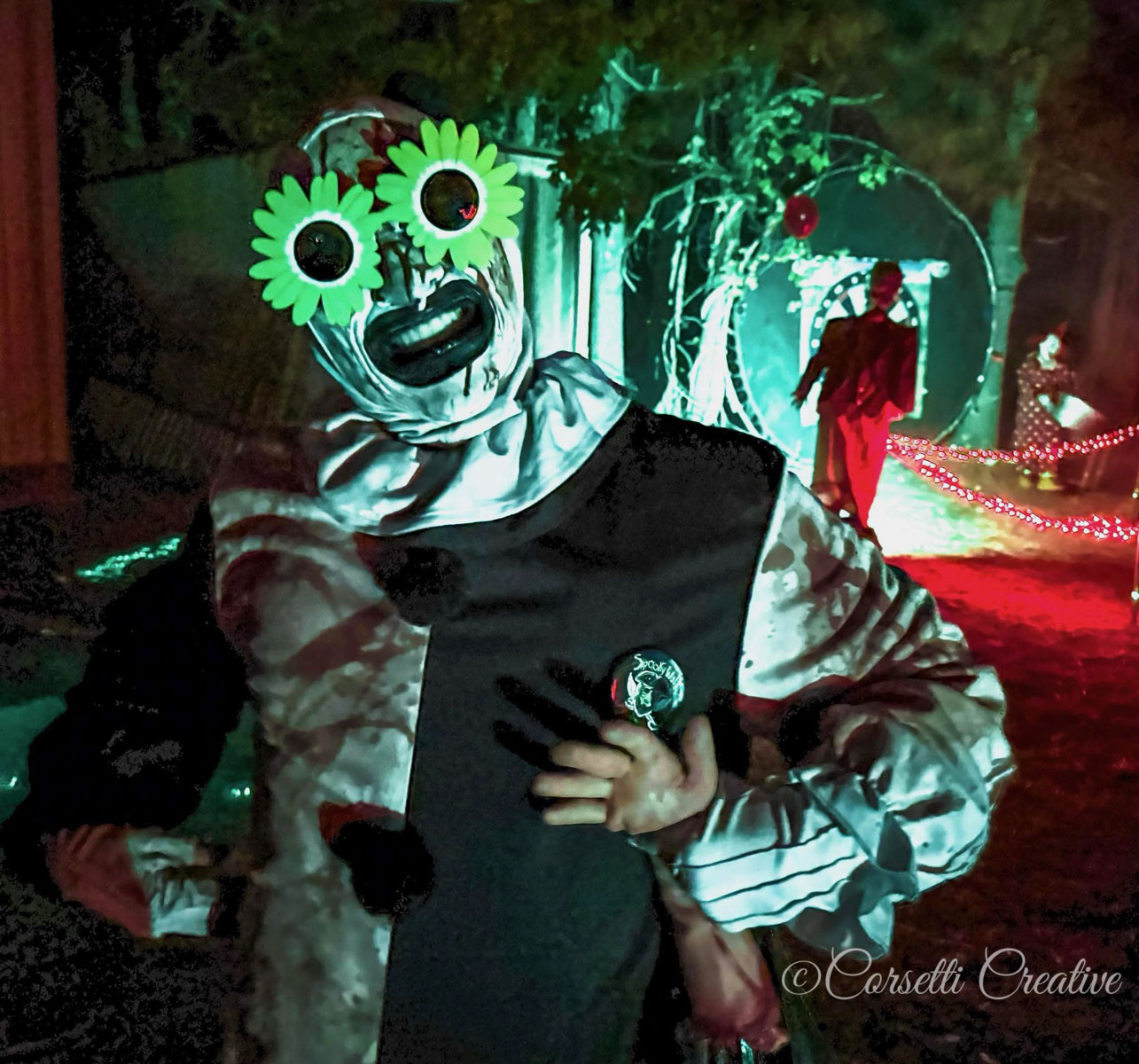 A sinister clown wearing oversized green flower-shaped eyes greets visitors along an eerily illuminated section of the Spooky Walk haunted trail. The approximately 45-minute experience features costumed performers, elaborate scenery and frightening encounters throughout the wooded grounds of Camp Pa-Qua-Tuck. Photo credit: Corsetti Creative, courtesy of Camp Pa-Qua-Tuck Fundraisers.
